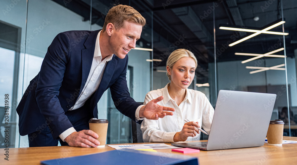 Fototapeta premium Professional male colleague mentoring or assisting a female coworker with a laptop in a sleek modern office environment.