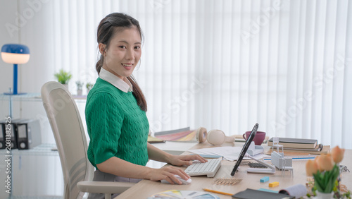A smiling interior designer sits at her desk typing on a keyboard while colorful design materials and furniture models surround her.