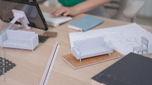 Two small scale sofa models sit on a wooden desk near a ruler and blueprint while a designer works with a tablet in the background.