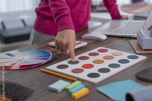 A designer's hand points to a green color swatch on a leather sample board next to a color wheel chart at her workstation.