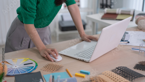 A close up of a designer's hand clicking a computer mouse beside an open laptop with a color wheel and woven samples on the desk.