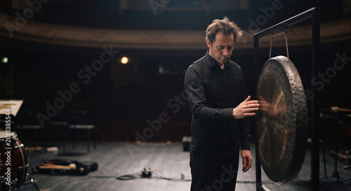 A candid, contemplative photograph of a percussionist standing alone on a silent, darkened stage after a powerful performance, resting a hand on the edge of the still-vibrating gong,