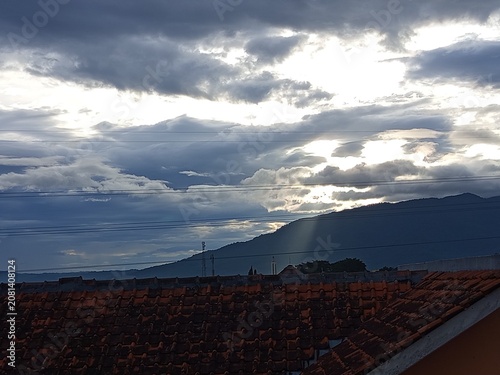 Dramatic Cloudy Sky with Sun Rays Over Red Tiled Rooftops and Mountain Silhouette