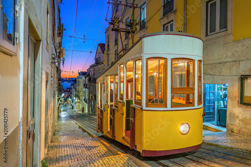 A classic yellow funicular tram ascending a steep, narrow cobblestone street lined with traditional colorful buildings  in Lisbon