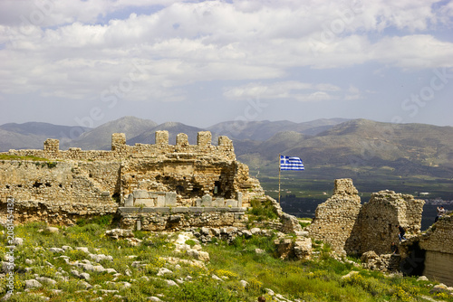 The ruins of a castle in Greece, with the Greek flag flying and mountains in the background.