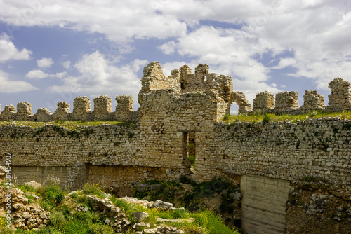 Ruins of a stone castle with crenellated walls and an arched opening under a cloudy blue sky.