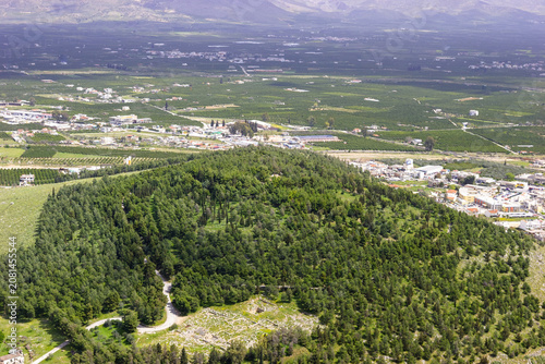 Aerial view of the ancient ruins of Mycenae, Greece, surrounded by lush green forests and agricultural fields, with the modern town visible in the distance.