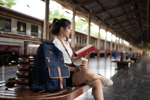 Asian woman traveler reading a book beside backpack while waiting calmly at railway station platform