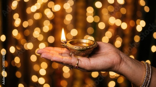 Woman holds traditional oil lamp with lit wick in hand against festive background lights