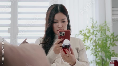 Asian woman holding drug bottle reading information on smartphone at home
