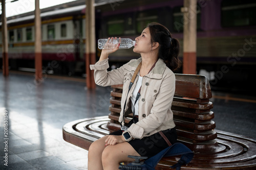 Asian woman traveler sitting on wooden bench and drinking water during a break at train station.