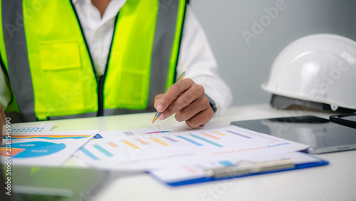 Фотография Engineer or project manager holding pen, analyzing yearly summary report chart and financial graphs on office desk with safety hard hat, managing construction project budget