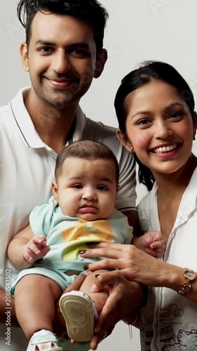 Indian young parents holding baby boy standing against white background, smiling and looking at camera, representing love, bonding, happiness, parenthood, family care and newborn lifestyle together