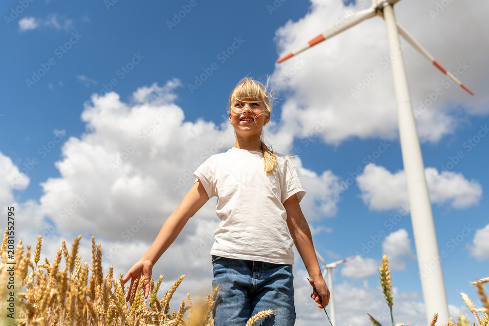 Fototapeta premium Smiling girl walks through golden wheat field under blue sky, with wind turbine and clouds showing clean energy theme. Rural summer scene connects childhood, harvest, nature, sustainability, freedom
