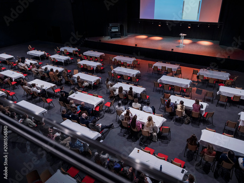 An elevated view of a conference audience seated at long tables before a stage in a large venue.