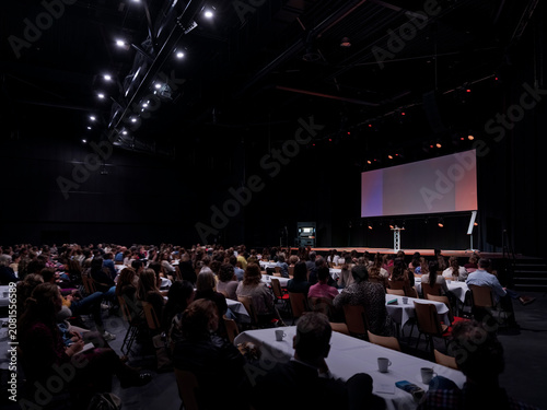 A crowd of attendees fills a big convention hall with a bright stage screen glowing in the dark.