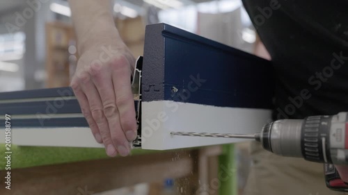 Close-up of a worker drilling into a black and white window frame profile during factory assembly, with hands and power tool visible