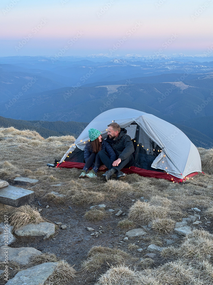 Fototapeta premium Happy couple sitting inside tent at mountain summit during sunset