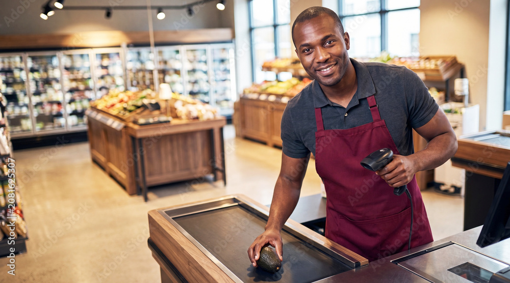 Obraz premium Smiling supermarket cashier scans a fresh avocado with a handheld barcode scanner at a modern grocery checkout.