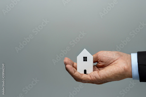 Close-up of a businessman's hand holding a small house against a gray background. Real estate, property investment, home insurance, mortgage loans concept. Copy space for text.