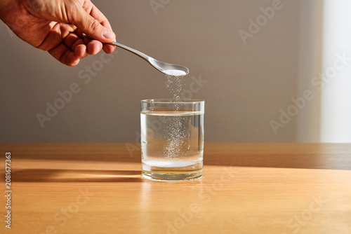 Fotografía Hand pouring white powder from spoon into a glass of water