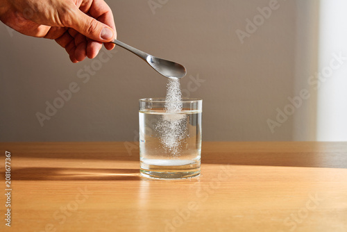 Bild auf Leinwand Hand Pouring White Powder from Spoon into Glass of Water