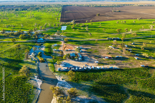 Aerial scene showcasing the Pilliga Artesian Bore Baths and adjacent campground, with a winding creek flowing through the lush green bushland and cultivated farmland stretching across the background