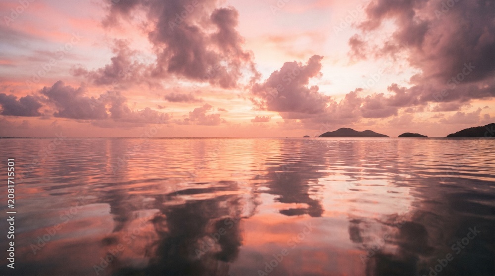 Obraz premium Sunset glow illuminates a calm ocean surface where soft pink clouds drift lazily above the distant horizon line reflecting vibrant colors across the gentle ripples of the peaceful tropical water.