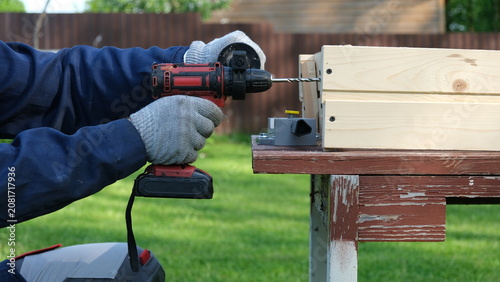 Photos A worker in a blue uniform and protective gloves drills a hole in a wooden structure with a cordless drill