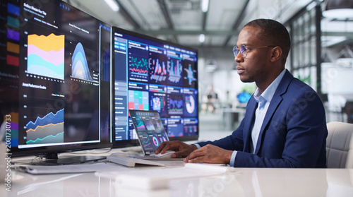 Black male data analyst with glasses working on multiple computer monitors showing charts and analytics dashboards at a white corporate office desk
