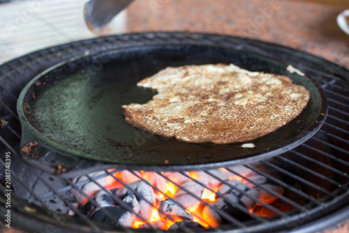 Traditional Pancake Prepared on a Cast Iron Griddle. A classic pancake cooks over live charcoal heat, creating a rustic outdoor breakfast scene.
