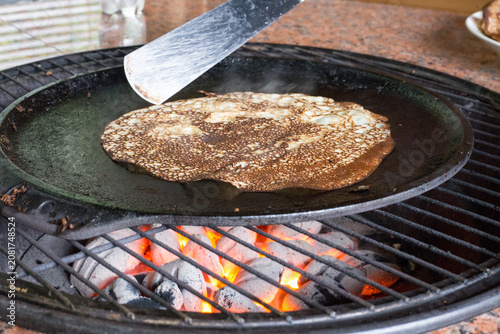 Rustic Outdoor Breakfast Prepared Over Live Charcoal Fire. Traditional pancake cooking on a cast iron griddle over glowing coals with visible heat and smoke.