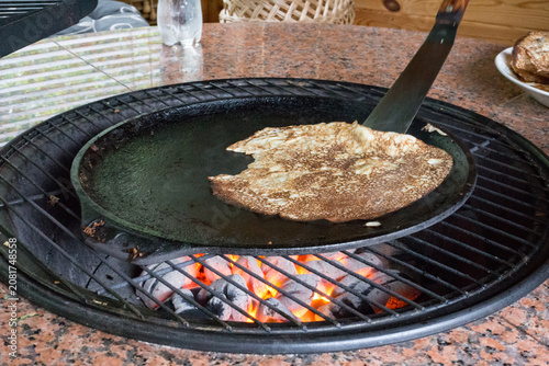 Homemade Pancake on Hot Cast Iron Pan Above Charcoal. Close-up of a pancake frying on a cast iron griddle with glowing barbecue briquettes underneath.
