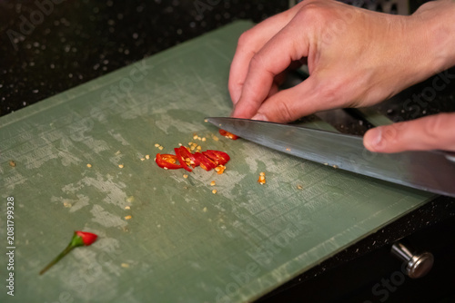 Man chef slicing fresh red chili pepper on green kitchen cutting board with knife. Culinary process of preparation vegetable for spicy dish. Healthy cooking and home food concept.