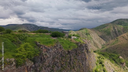 Exploring the  Garni ancient temple  in Armenia on a cloudy day surrounded by mountains. Visitors walk near an ancient temple located on a cliff in Armenia. The sky is cloudy and the mountains are vis