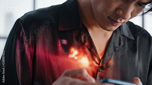 Young Asian man intensely playing a mobile game on his smartphone. Colorful reflection of the action game is projected on his shirt. Modern digital entertainment and esports concept