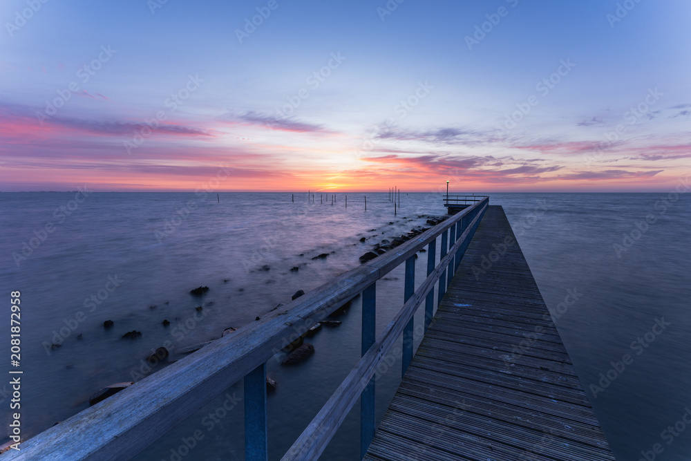 Obraz premium Landscape photograph of a wooden pier stretching into Markermeer near Volendam in North Holland, Netherlands, during a peaceful sunrise. The first light of day paints the sky in soft shades.