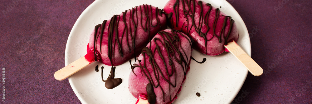 Fototapeta premium Three popsicles made from strawberries, bananas, beets, and Greek yogurt sit on a plate. They are drizzled with chocolate and enjoyed during a sunny day with friends, banner