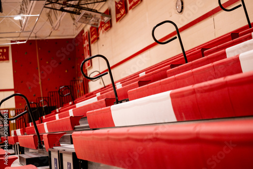 Фотография Rows of empty red tiered bleacher seating with black safety handrails extend across a school gymnasium, photographed at an angle