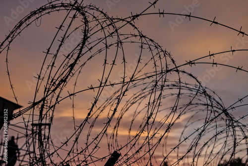 Obraz na plátně Barbed wire coils silhouetted against a glowing sunset, evoking freedom, captivity and hope