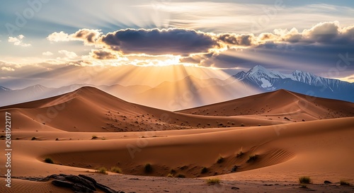 Golden Sunbeams Breaking Through Clouds Over Vast Desert Sand Dunes With Snow Capped Mountain Peaks At Sunset