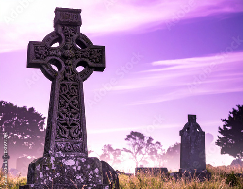 An intricate stone Celtic high cross stands silhouetted against a purple sunset in a quiet, historic graveyard in Meath.
