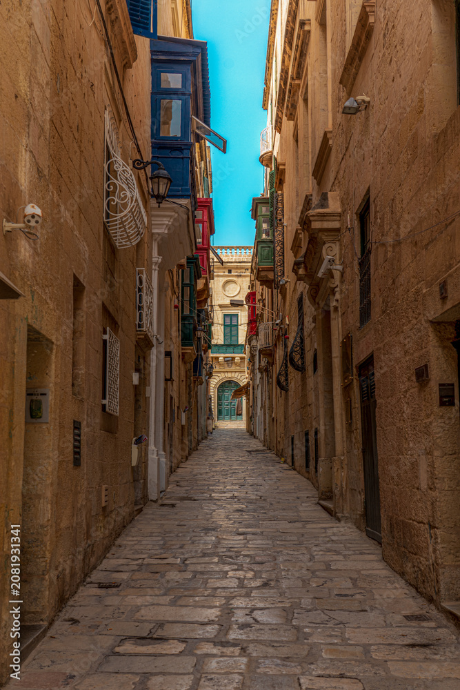 Fototapeta premium Narrow Alleyway road in valletta, malta with limestone buildings lining each side with colorful balconies and windows and clear blue summer skies overhead
