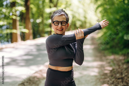 Mature woman in black athletic wear stretches her arms while standing on a tree-lined path in a park during daylight, showcasing a healthy lifestyle and fitness routine