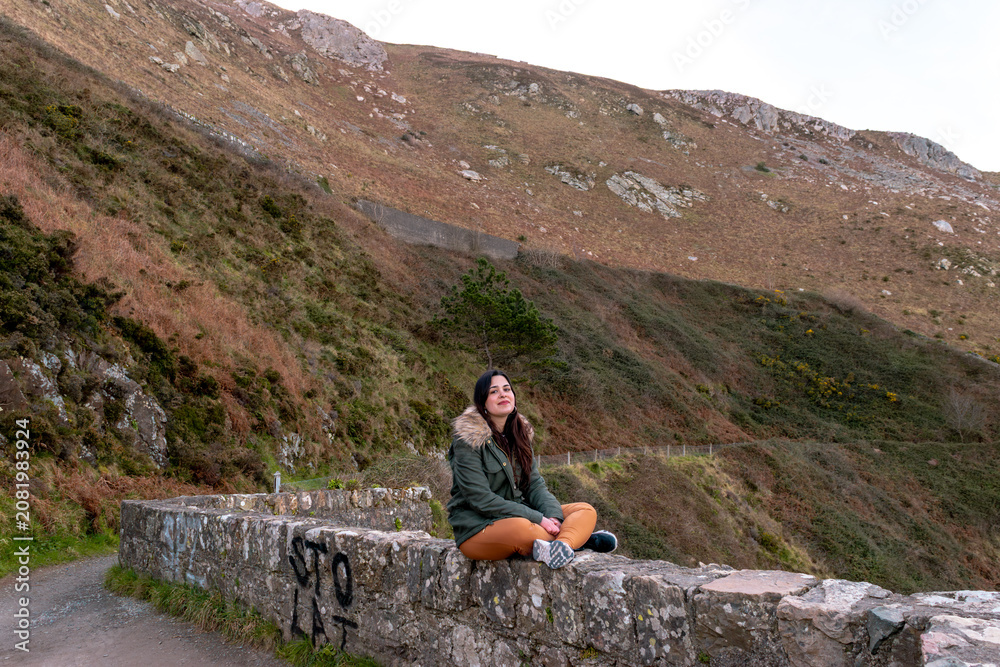 Fototapeta premium Young woman sitting cross-legged on a stone wall along a mountain trail in Ireland