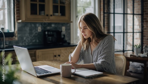 Young woman focused on her laptop while working from home in a cozy kitchen setting