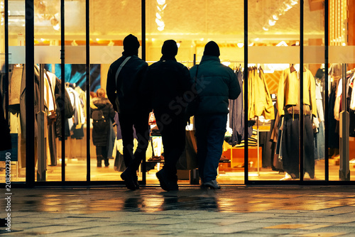 Obraz na plátně Silhouettes of three men entering a brightly lit fashion clothing store at night