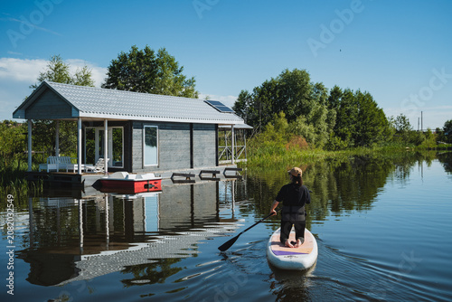 Φωτογραφία Person kneeling on paddleboard moving toward rustic boathouse beside calm reflec