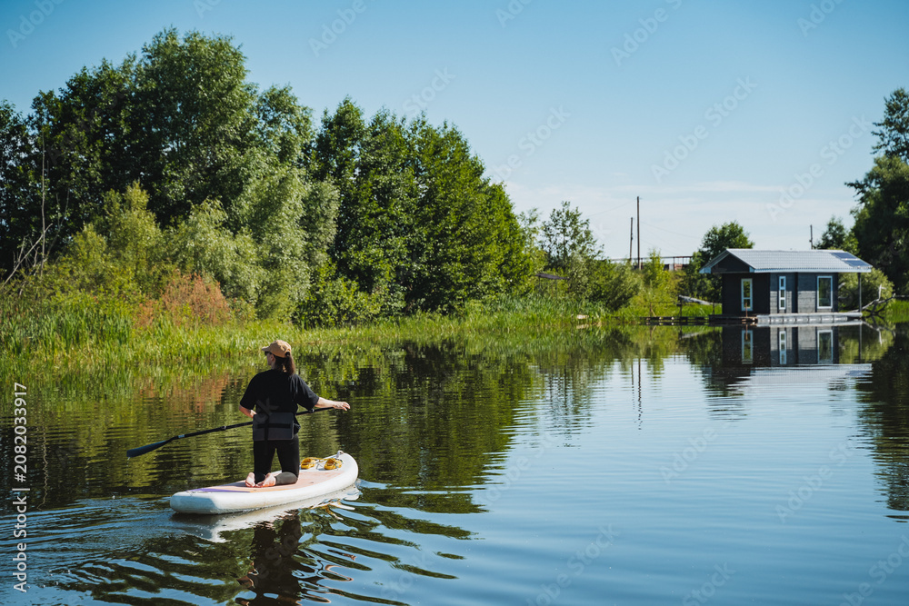 Fototapeta premium Person paddling standup paddleboard along calm river beside tree lined shore, reeds, reflections, and small docked cabin in peaceful summer landscape under bright daylight