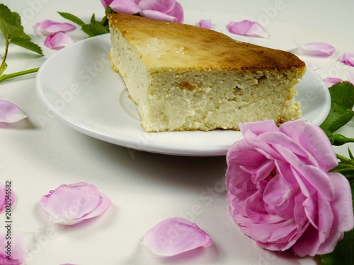 A white plate with a piece of homemade cottage cheese pie on it stands on a white table among roses and petals, side view
